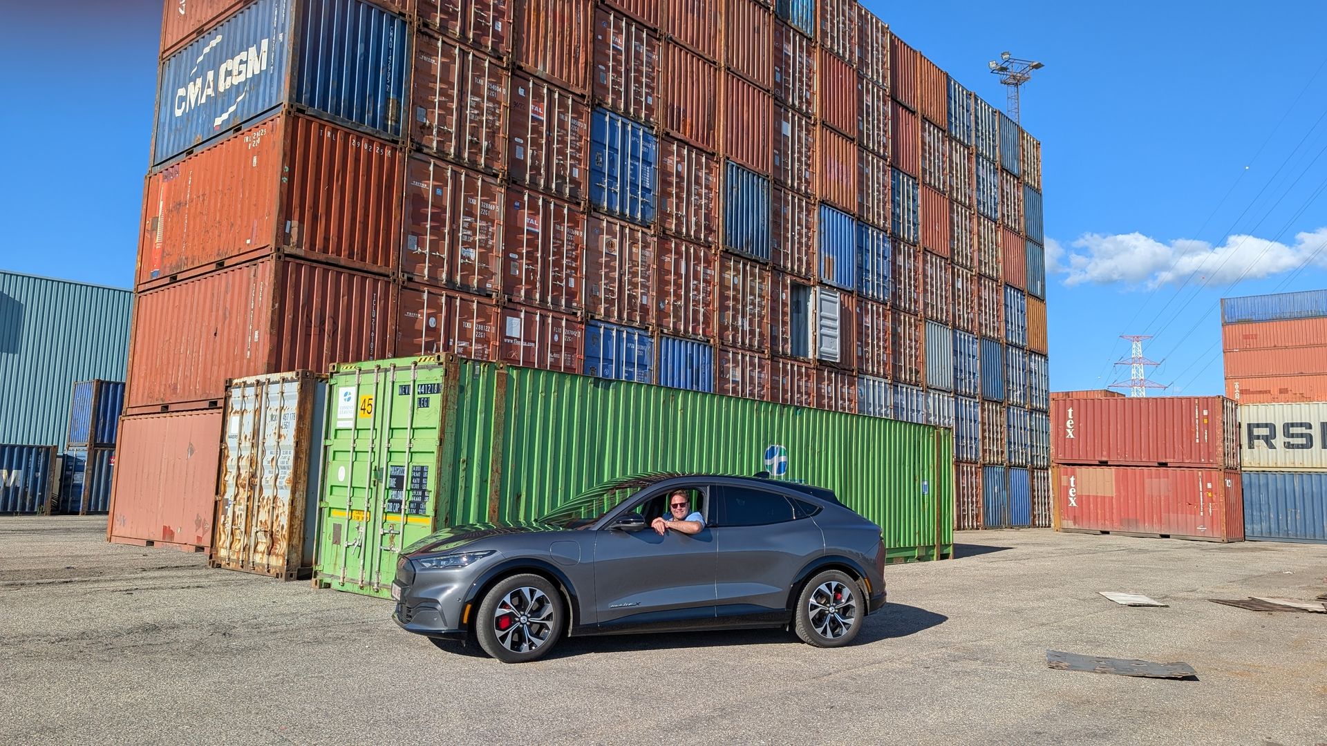 Gray SUV in a cargo container yard, person in driver's seat, blue sky, stacked containers in the background.