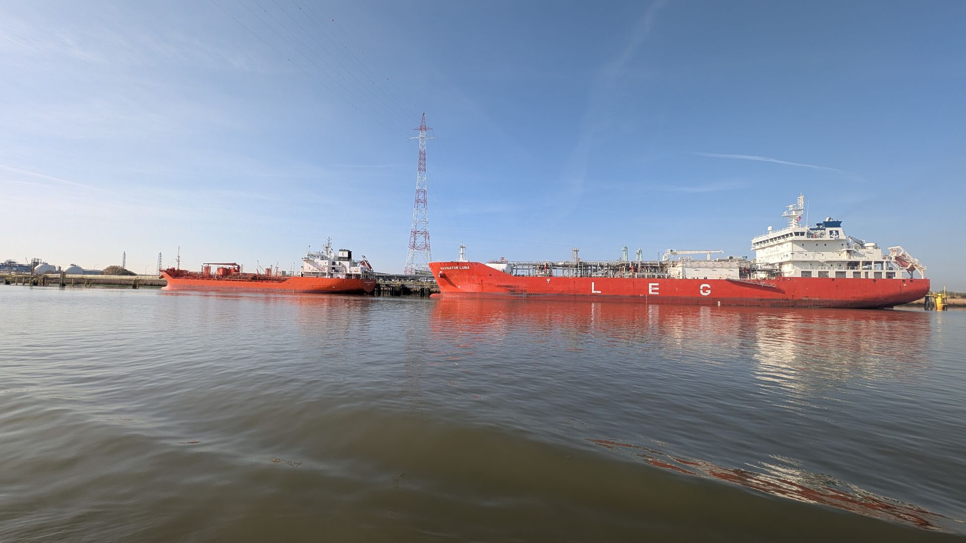 Red cargo ships docked on calm water under a clear blue sky.