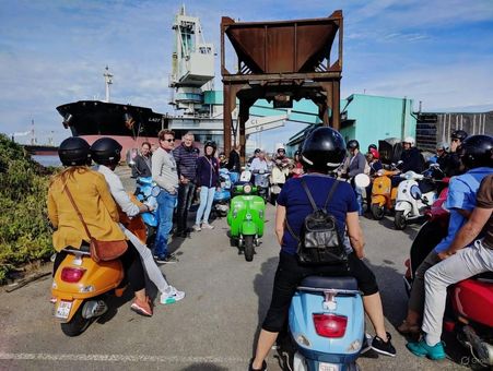 A group of people on colorful scooters, gathered near a harbor with a large ship and building in the background.