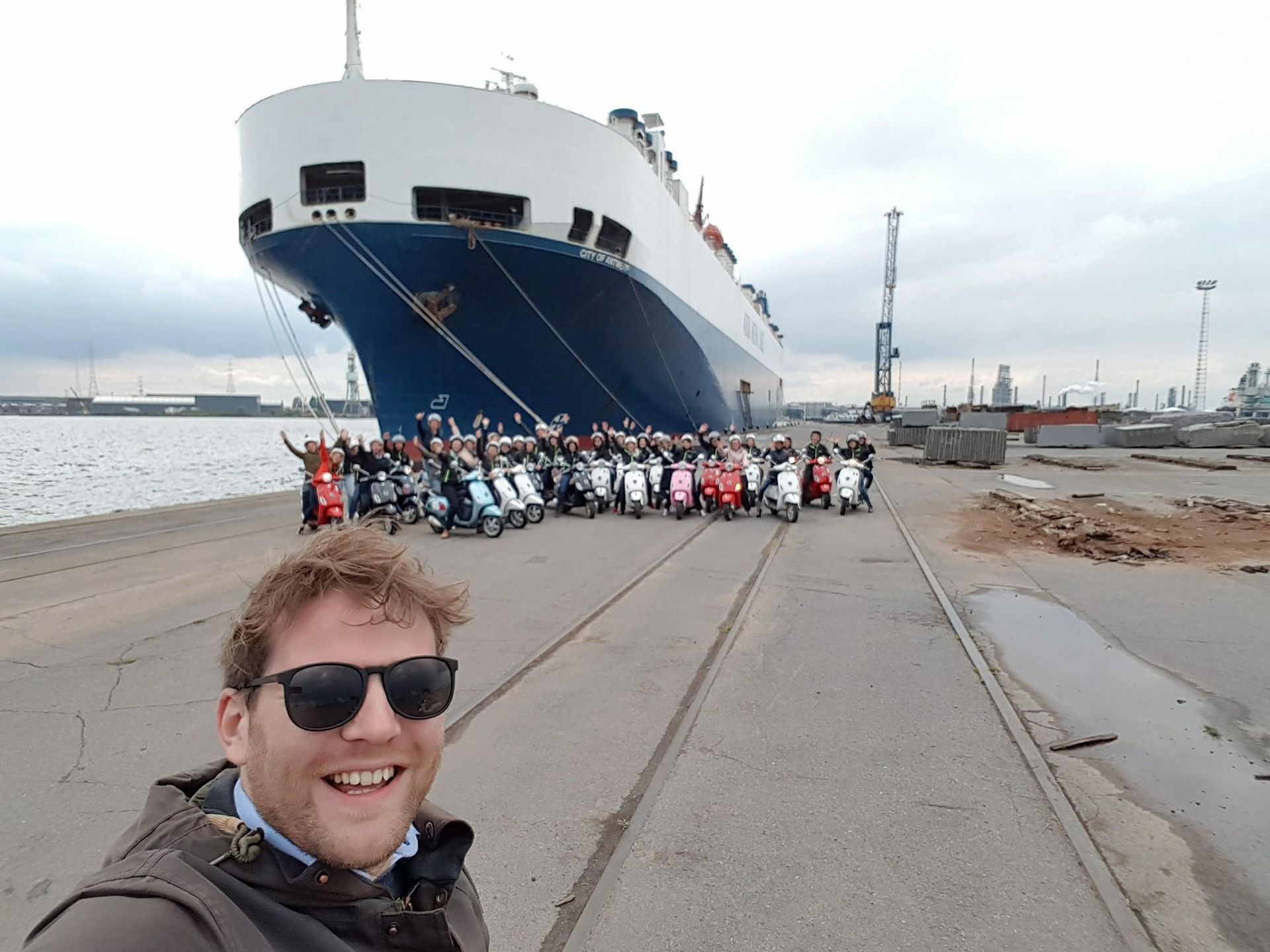 Man takes selfie with group of scooter riders in front of ship at a port.