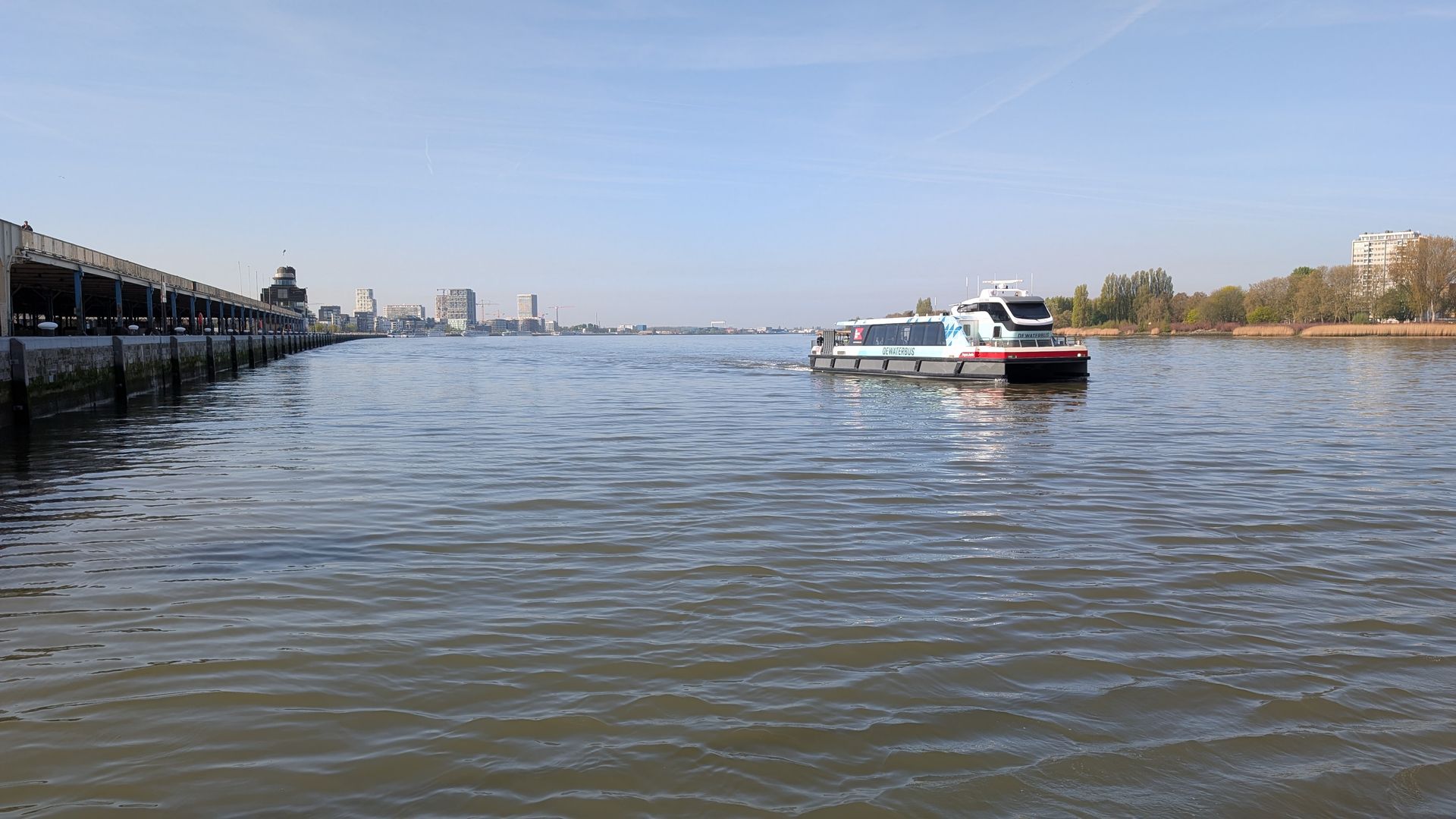 River with a ferry and boats under a clear sky, with a pier on the left and shoreline in the distance