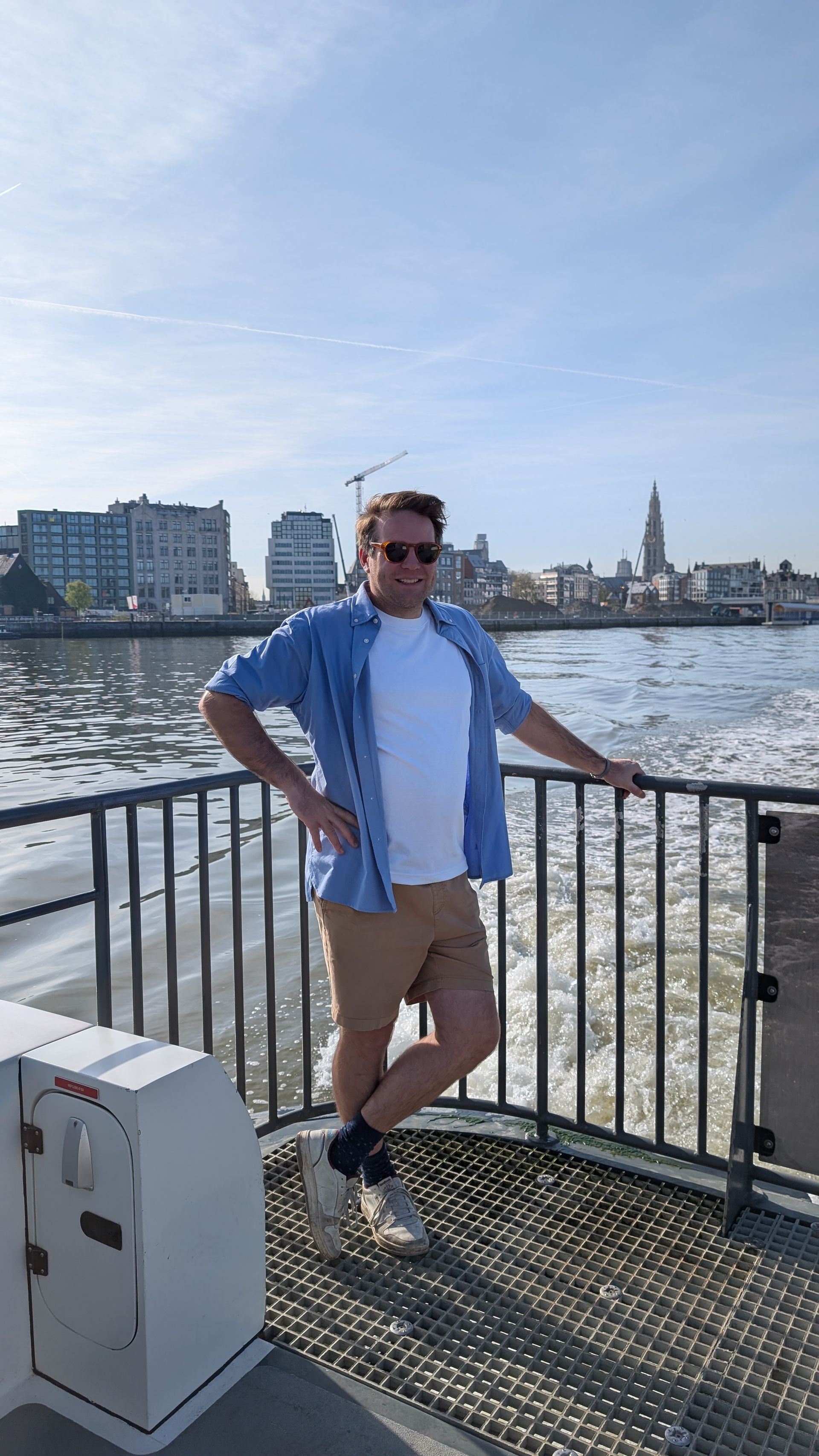 Man leaning on a railing by the water, with city buildings and a church spire in the background.