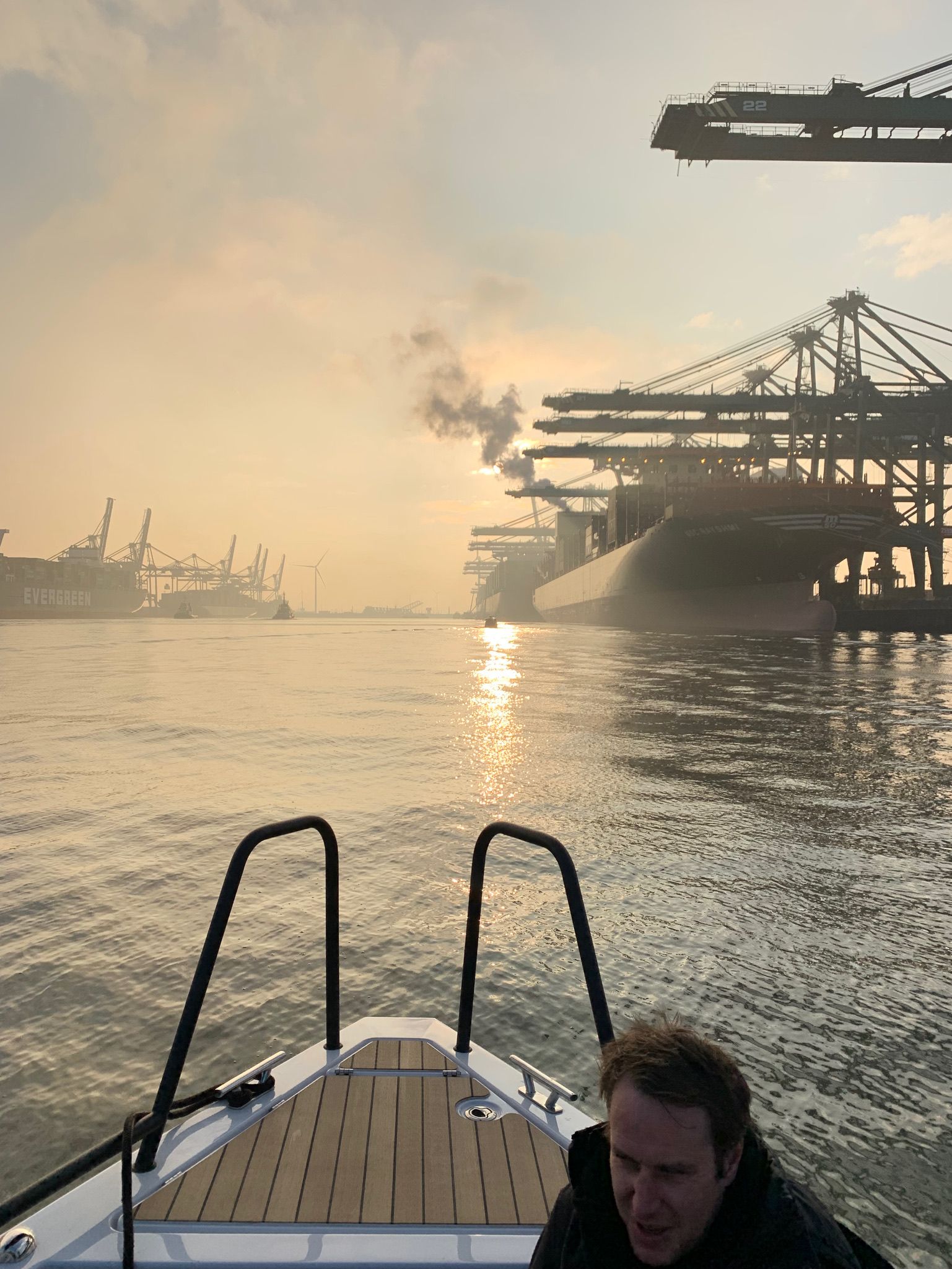 Boot fährt bei Sonnenuntergang in Richtung Hafen. Mann im Boot, goldenes Licht, Kräne und Schiffe.