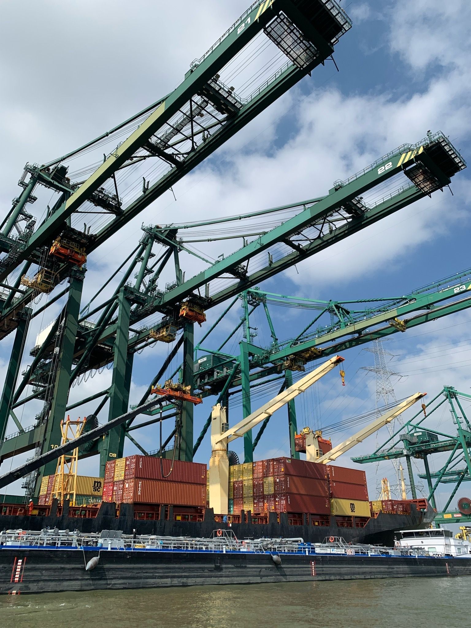 Große grüne Kräne beladen Frachtschiffe in einem Hafen unter einem teilweise bewölkten Himmel.
