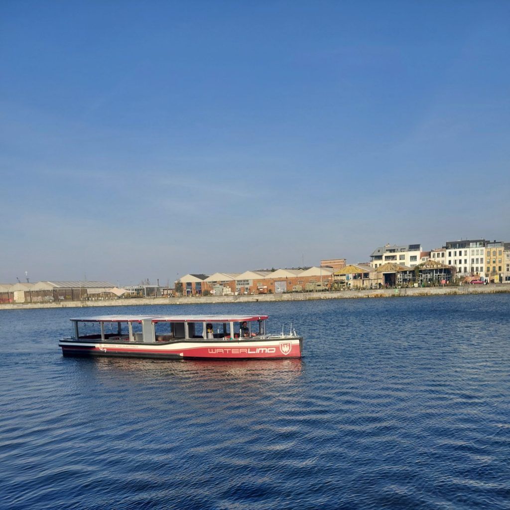 Red boat on blue water near a waterfront lined with buildings under a clear sky