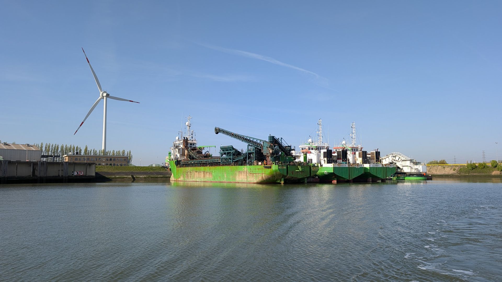 Large green fishing boats on calm water near a wind turbine under a clear sky