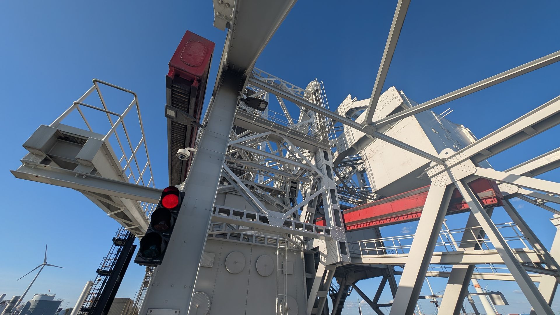 Low-angle view of a large industrial structure with red accents, a traffic light, and a blue sky.