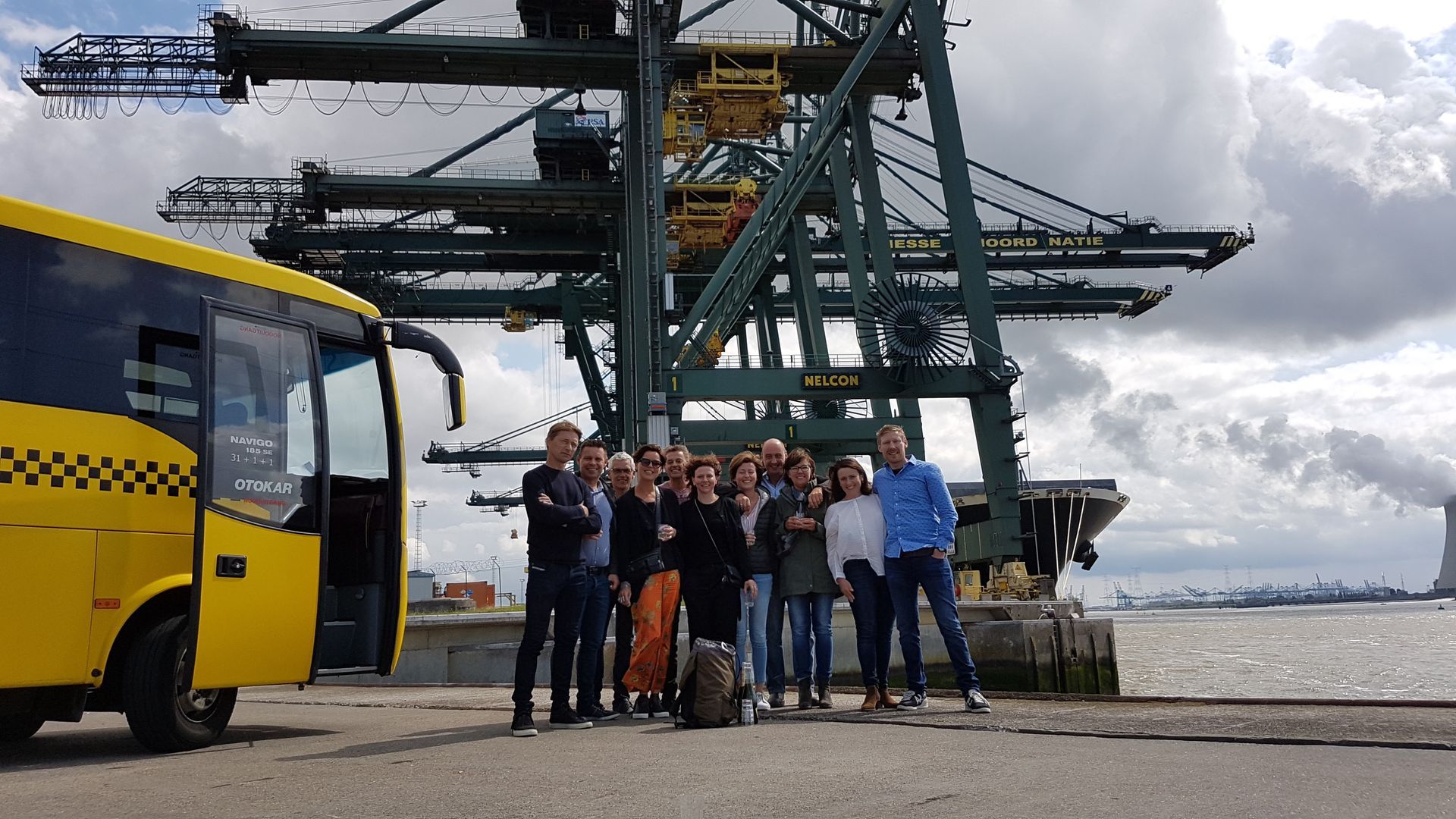 Group of people posing by yellow bus and large crane at a port. Cloudy sky.