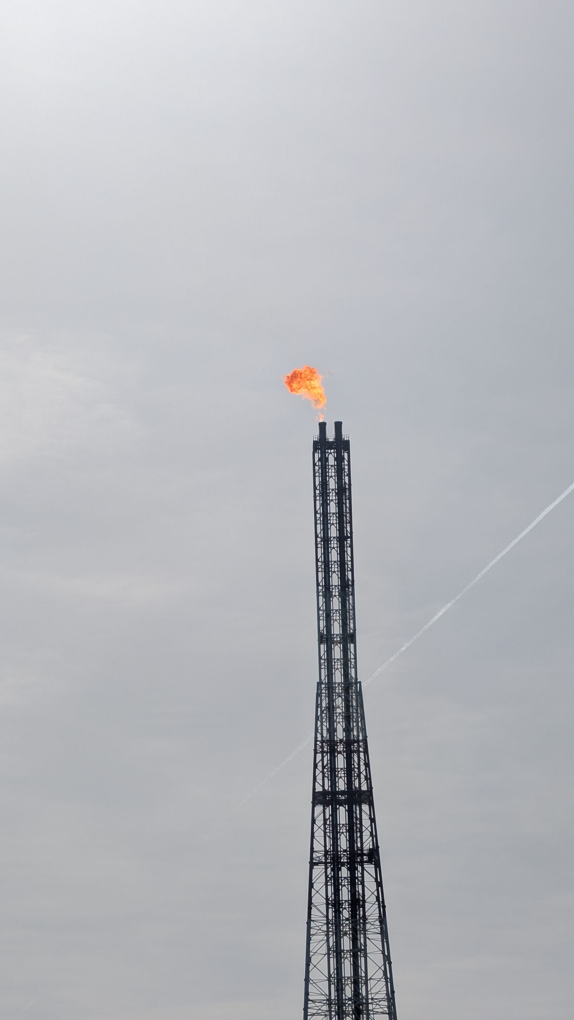 Tall industrial flare stack burning orange against a gray sky