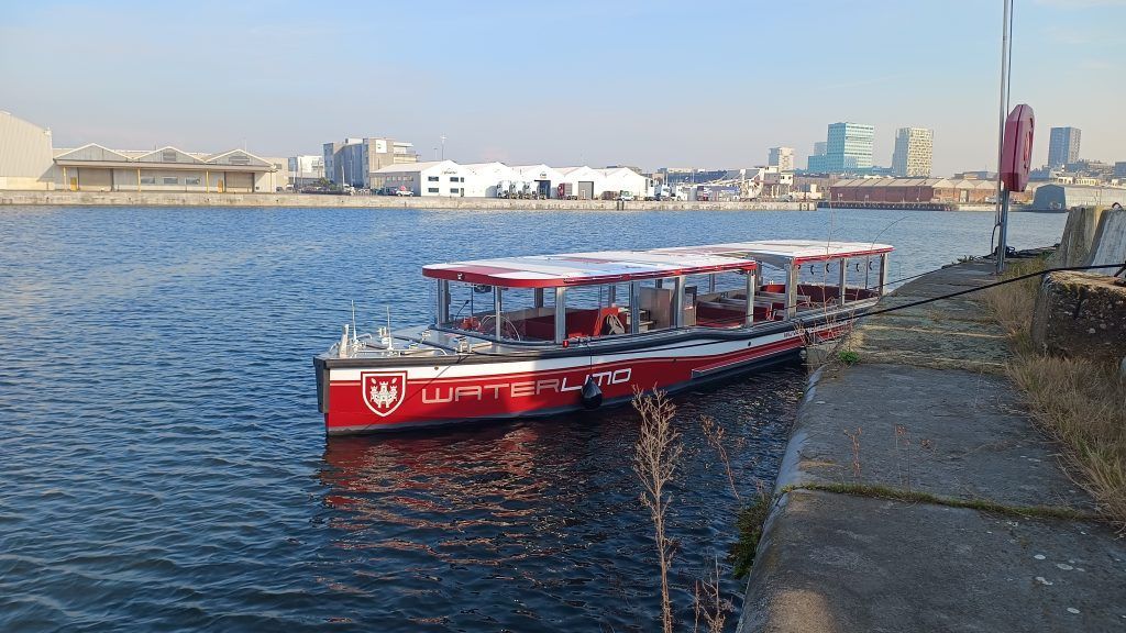 Red river ferry docked by a stone embankment, with city buildings across the water.