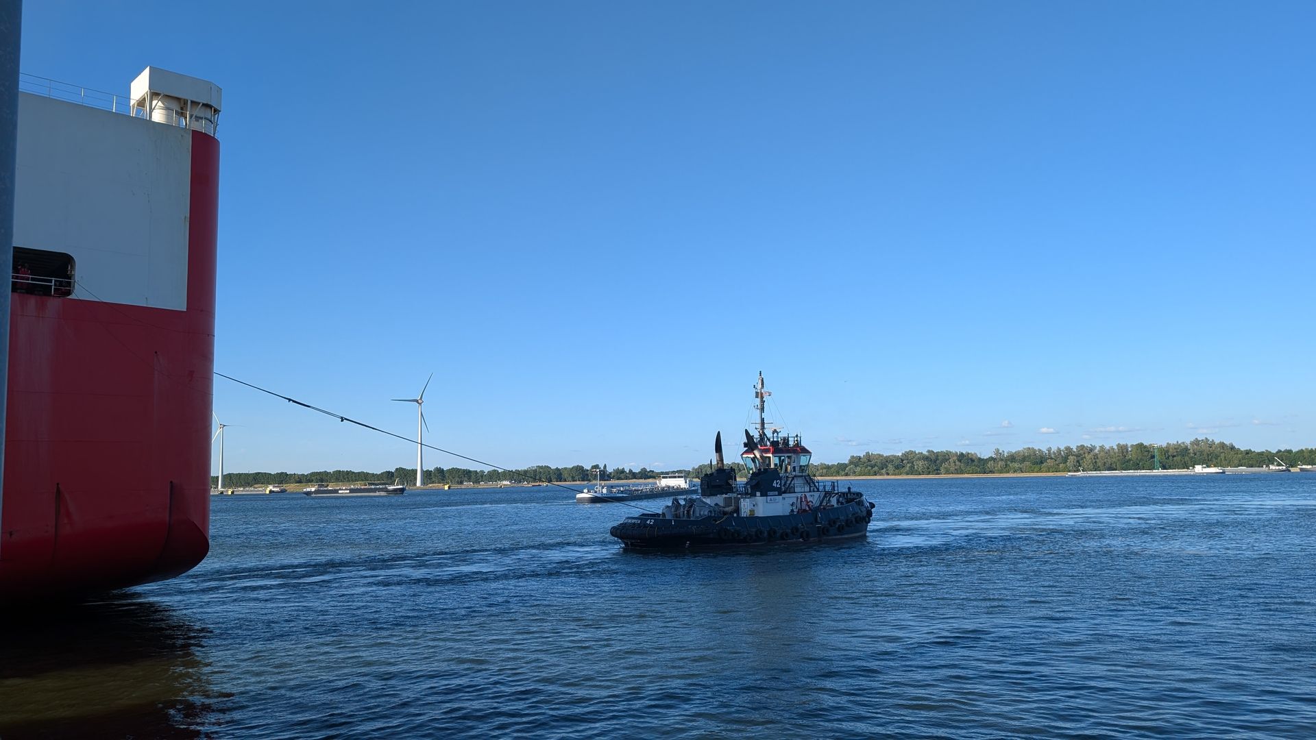 Schlepper hilft einem großen rot-weißen Schiff in einem Hafen unter blauem Himmel.