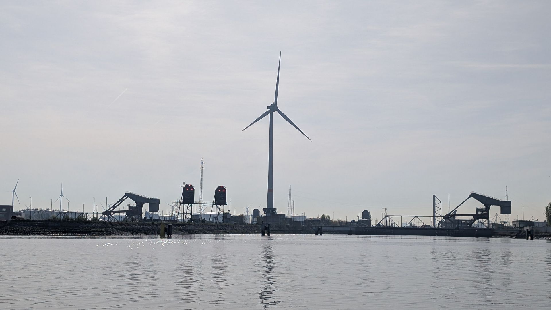 Industrial waterfront with a wind turbine, cranes, and calm water under an overcast sky