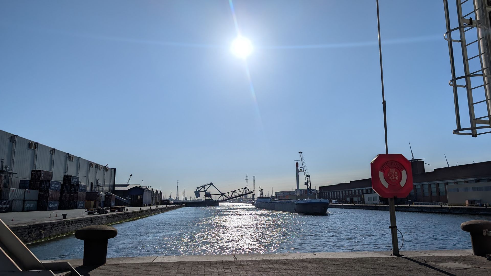 Een zonnige dag in een haven met een schip, kranen en gebouwen. Rechts een reddingsboei.
