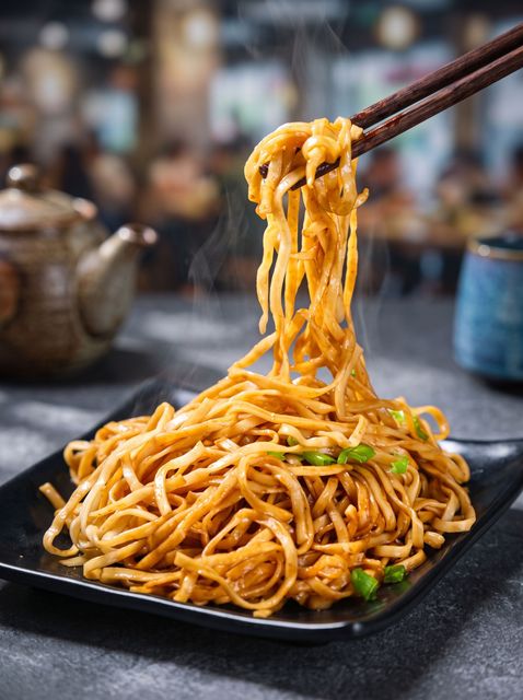 Chopsticks lifting saucy noodles from a black plate, with a blurred restaurant background