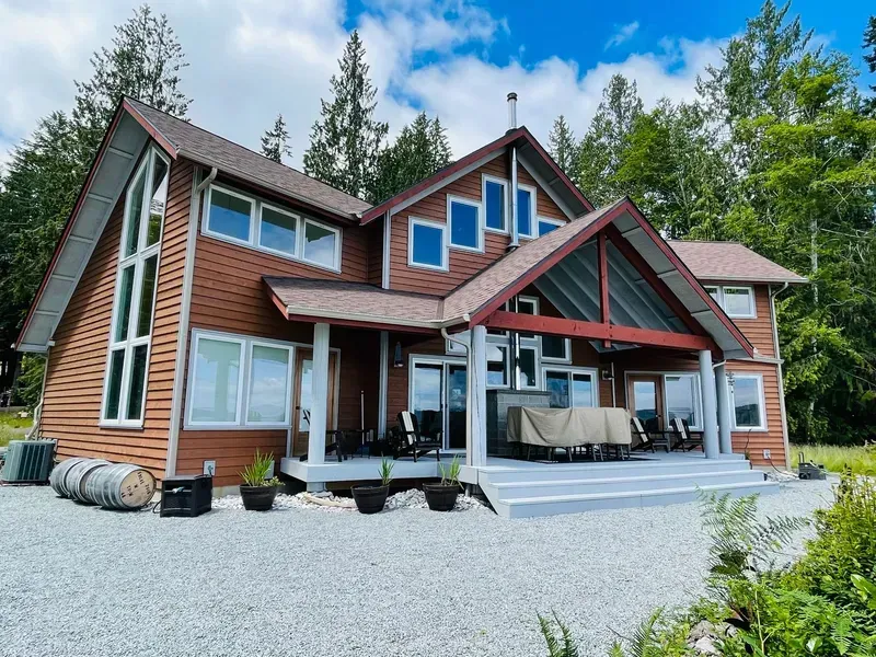 Large red-brown cabin-style house with steep roofs, white trim, and a gravel driveway under a blue sky