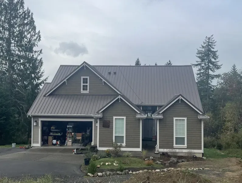 Gray house with attached garage and metal roof, surrounded by trees on a cloudy day