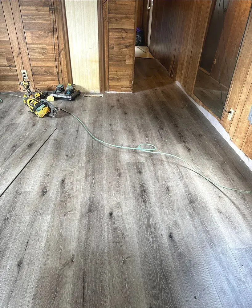 Wood-paneled room with gray flooring being installed, tools and extension cord on the floor.