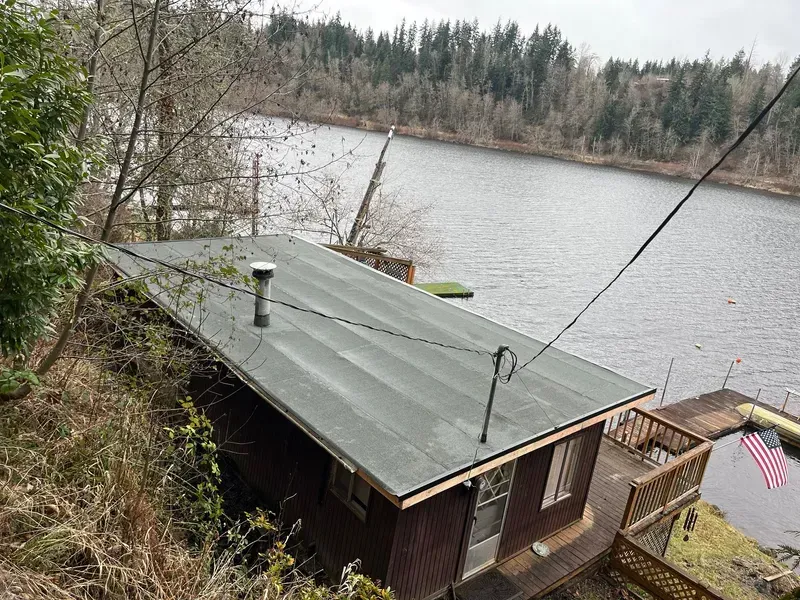 Small cabin with green roof beside a lake, surrounded by trees and a dock with an American flag.