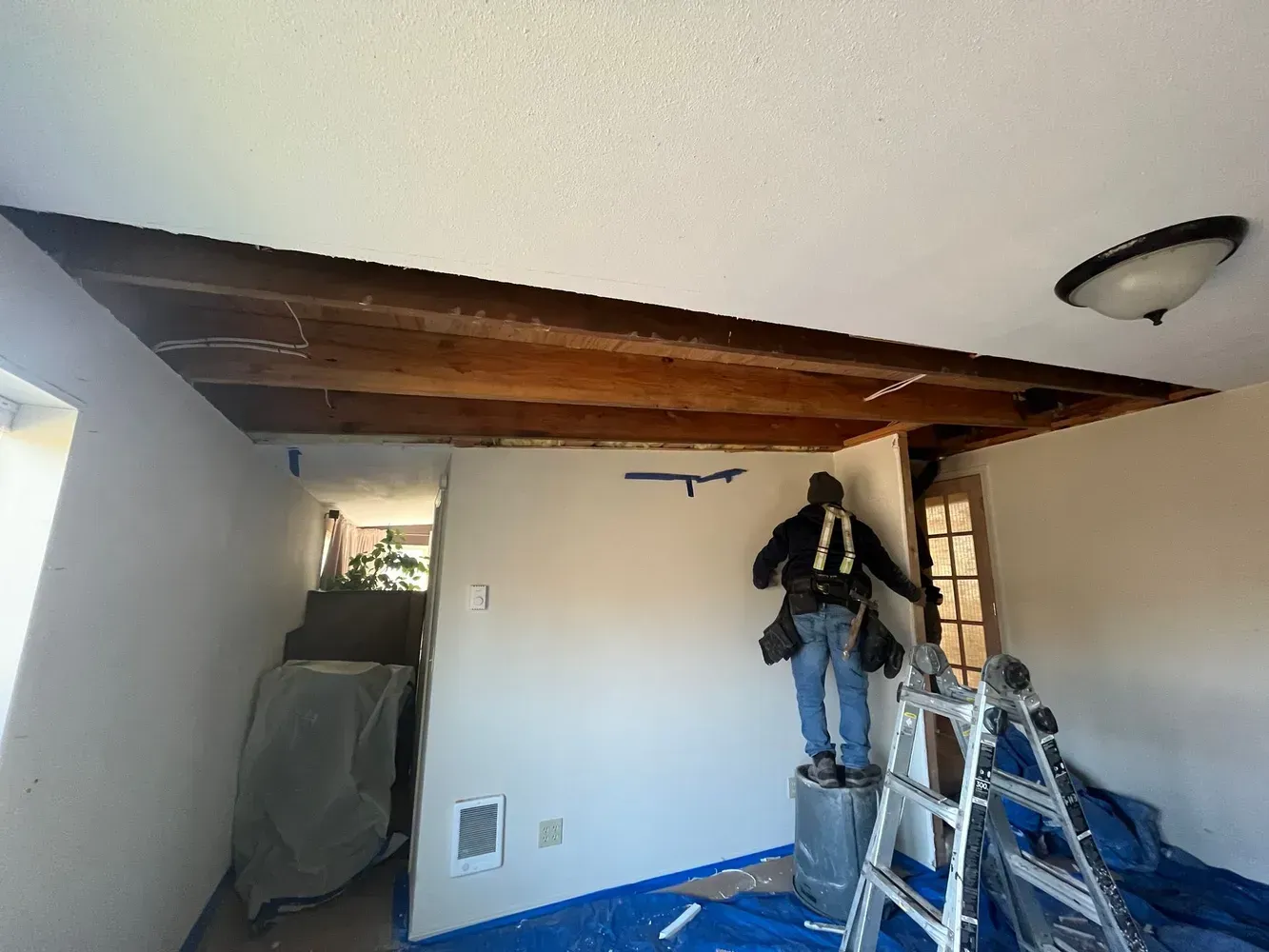 Low-ceiling room with exposed wood beams, ladder, hanging clothes, and covered furniture in renovation clutter