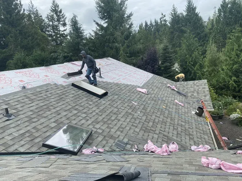 Workers repairing a gray shingled roof on a house surrounded by trees