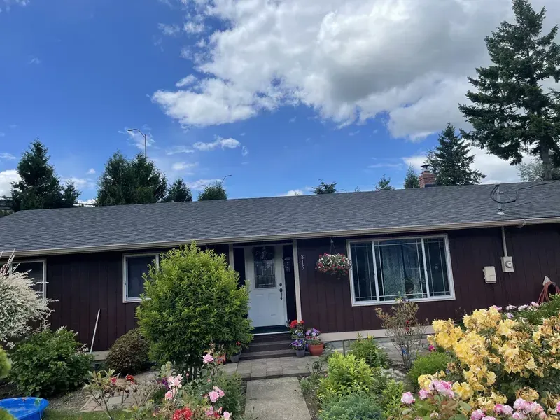 Single-story house with dark siding, white door, and flowered front garden under a blue, cloudy sky