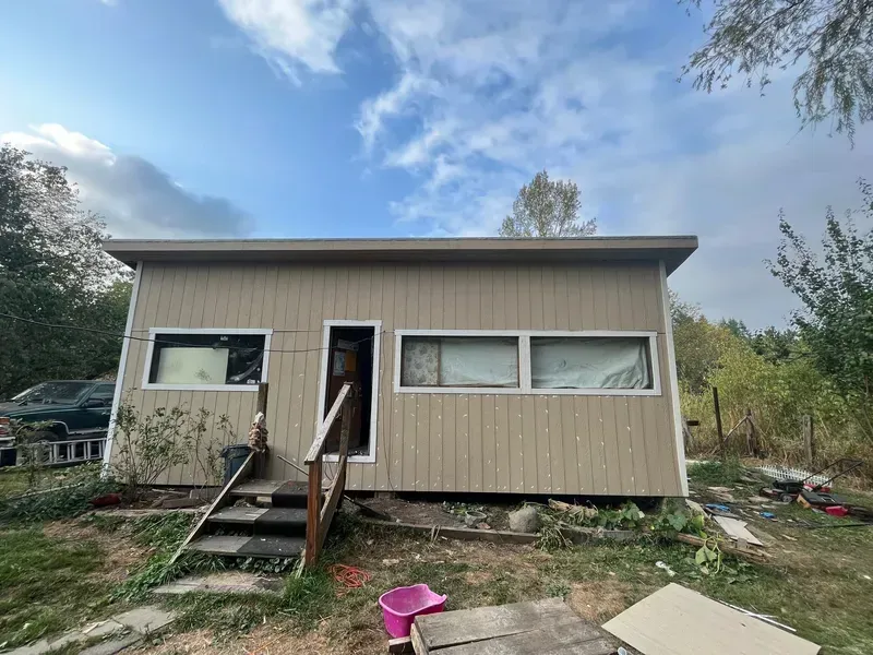 Small beige cabin with front steps and open door, set in a grassy yard under a cloudy sky.