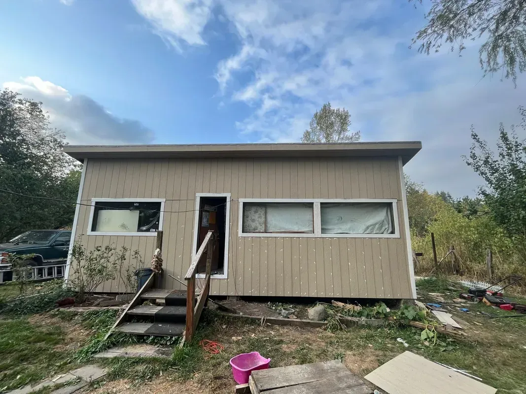 Small beige house with damaged front steps and overgrown yard under a cloudy sky
