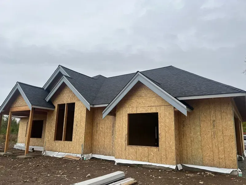 Partially built single-story house with tan wood siding and dark gray roof under cloudy sky