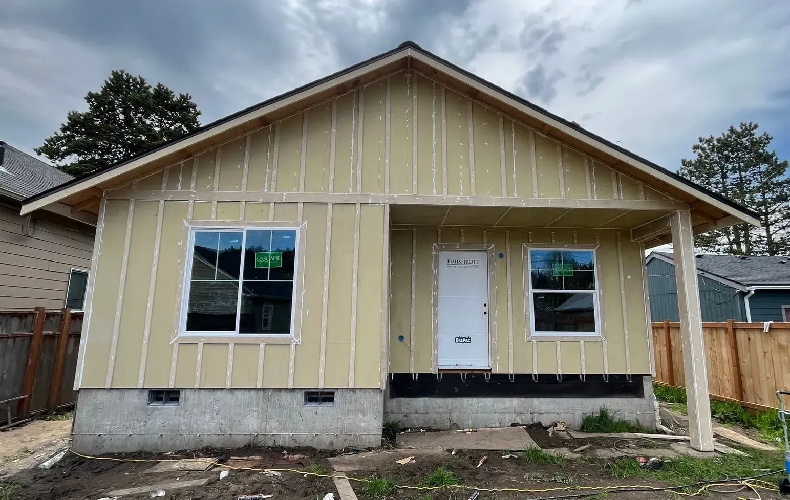 Front view of a small tan house with white trim, white door, and concrete foundation under construction.