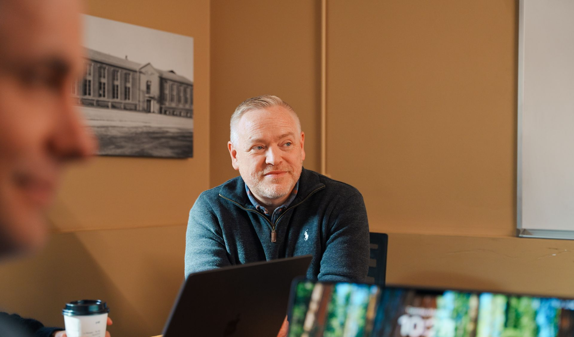 Scott Linklater in a meeting, looking towards the left, wearing a dark sweater. Beige wall behind him with a framed picture.