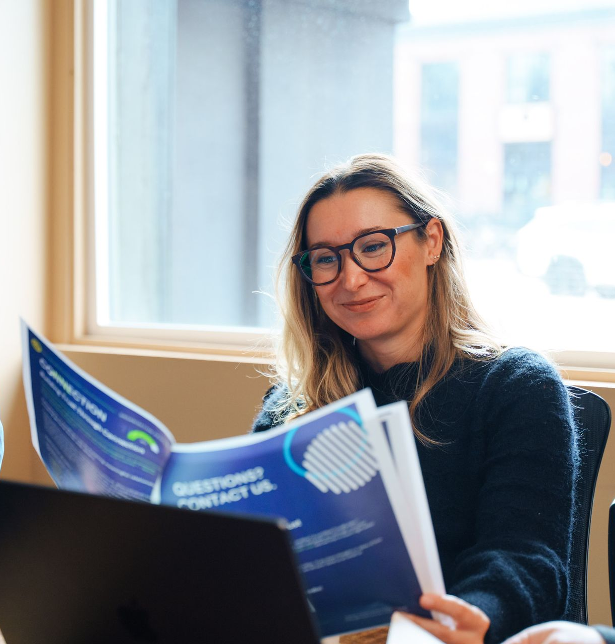 Woman with glasses smiles while reading a brochure near a window and laptop.
