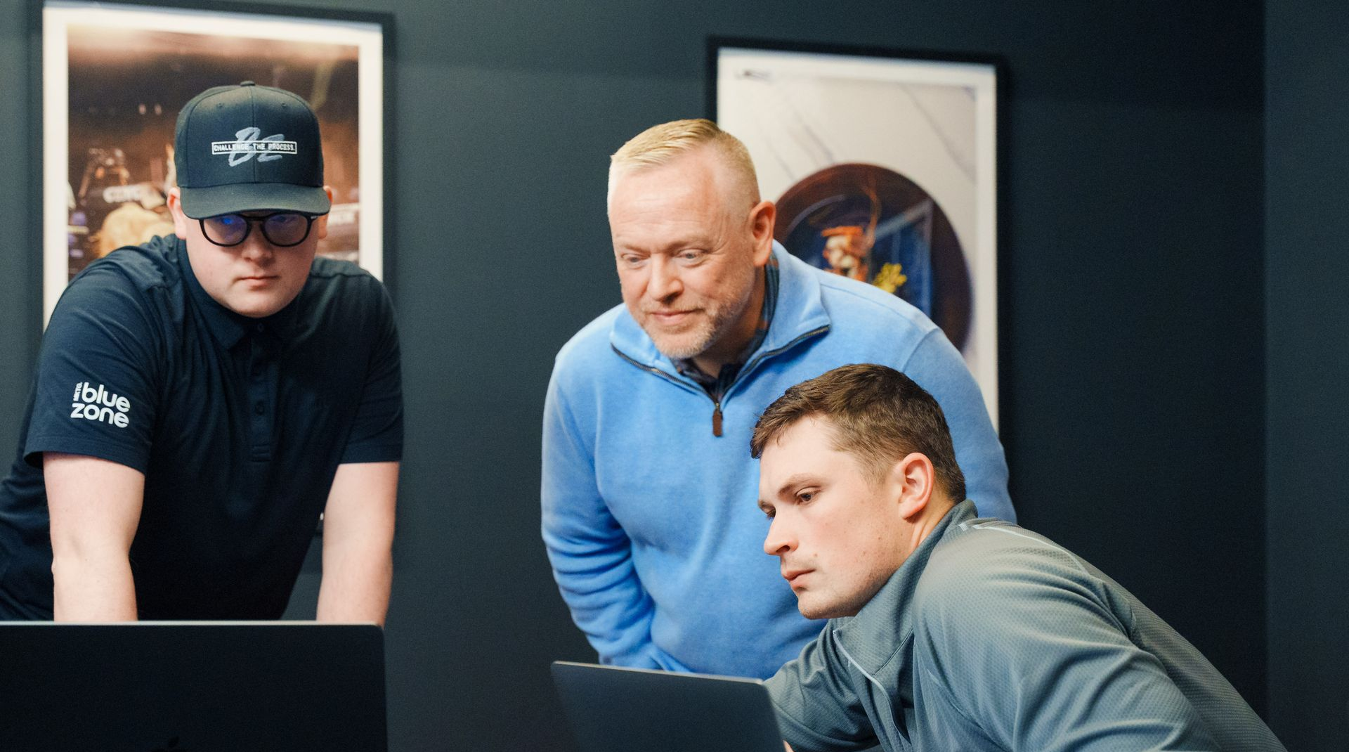 Three men looking at laptop screens in a dark room. One wears a hat, another a blue sweater, and the third a gray jacket.