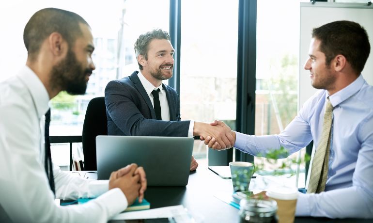 Three men sitting at a conference table, two are meeting and shaking hands