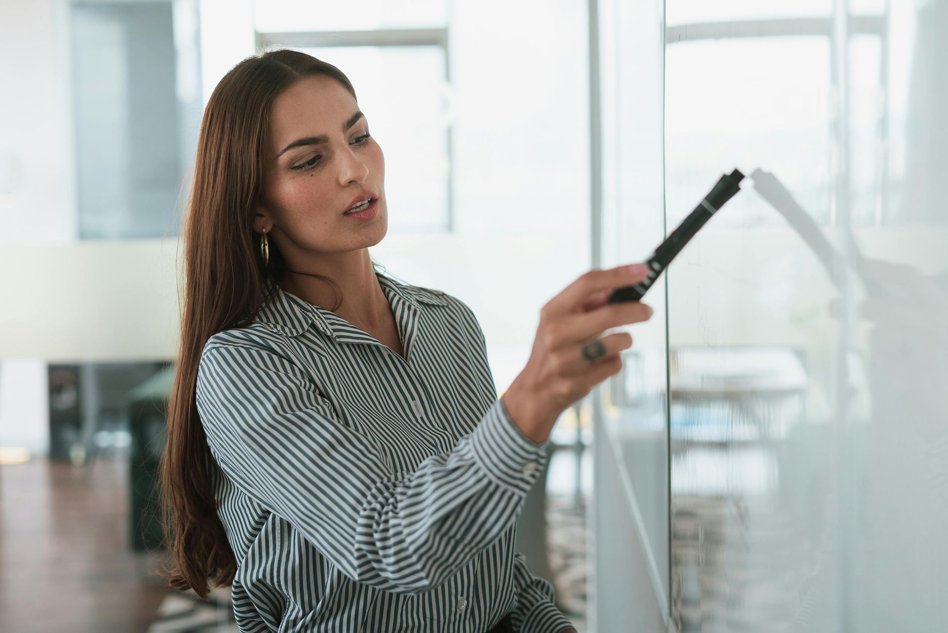 Woman in striped shirt pointing at a whiteboard with a marker; indoors.