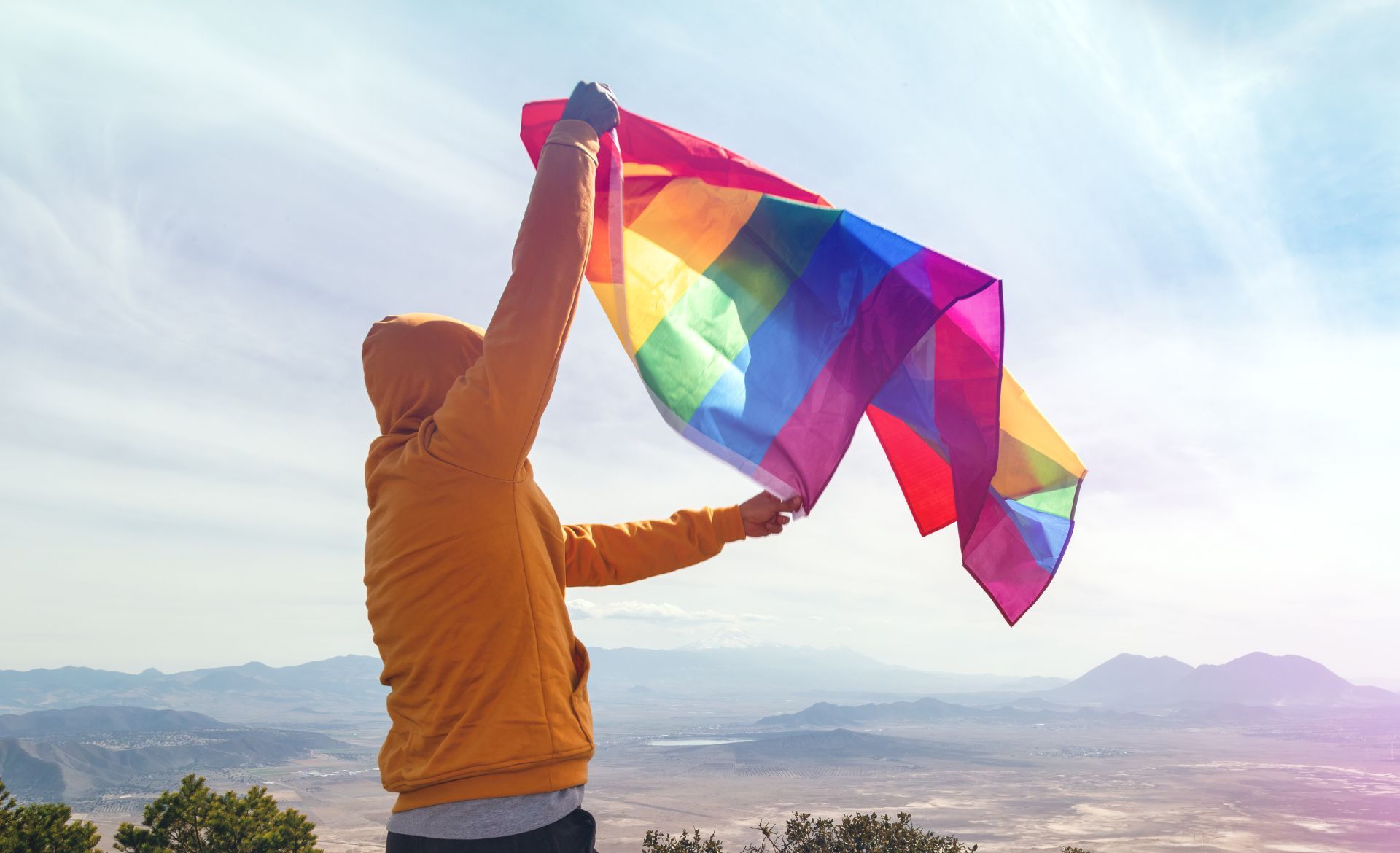 Person in orange hoodie holds up a rainbow flag on a mountain, sunny day.