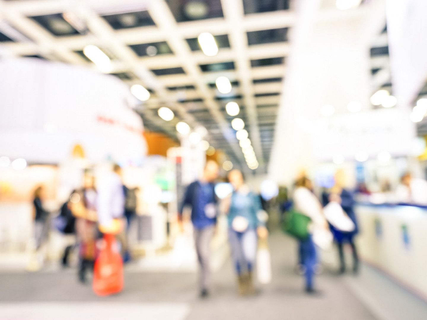 Blurred view of people in a large, brightly lit convention hall; a few figures walk toward the camera.