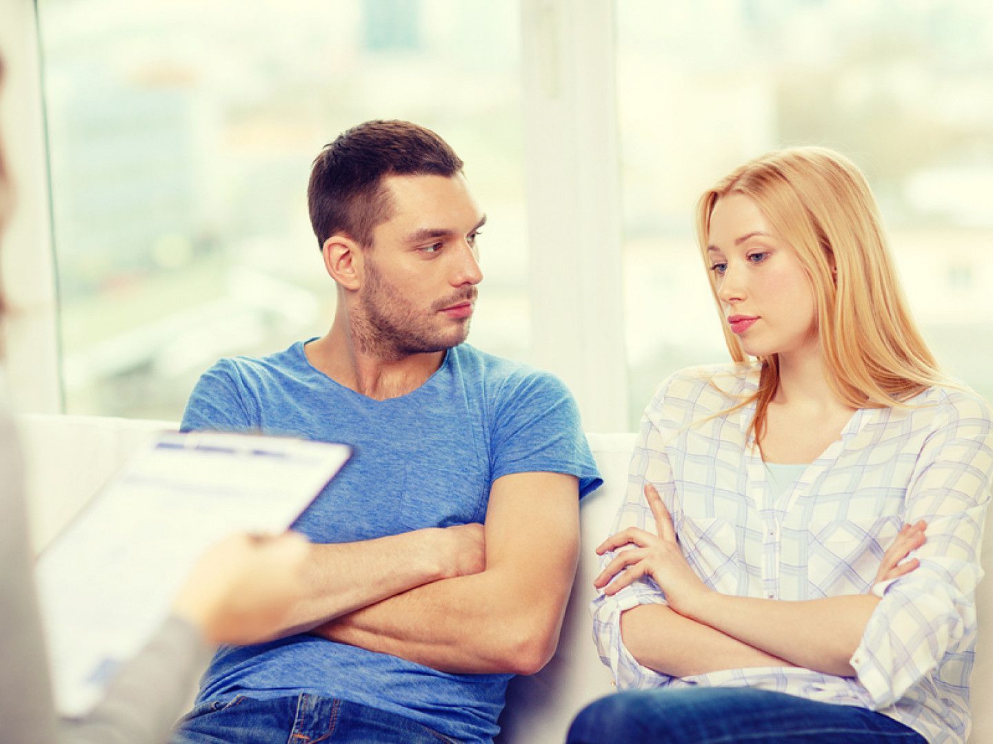 Couple with arms crossed, looking at therapist holding clipboard, in an office setting.