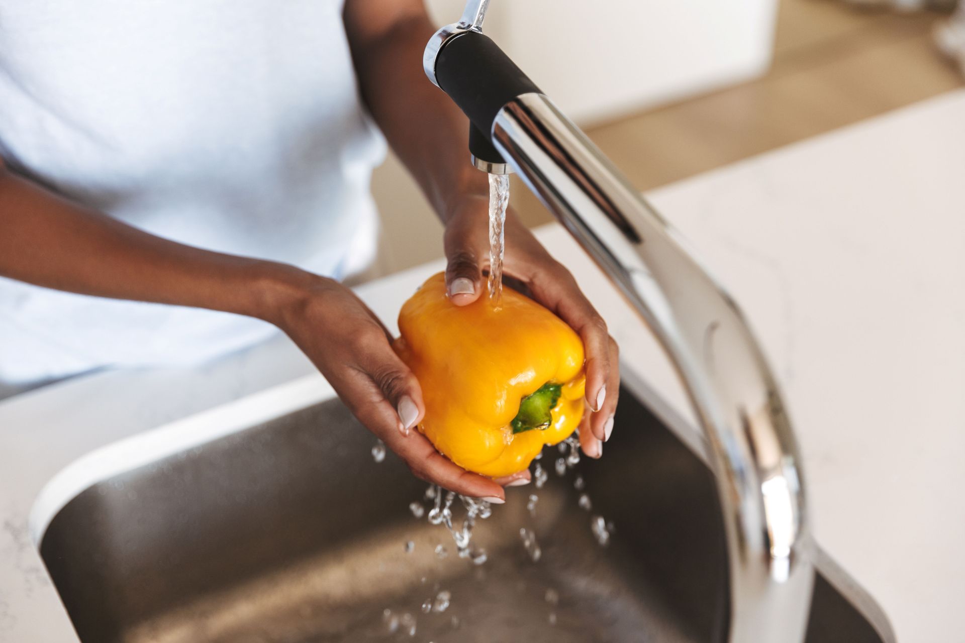 Hands rinsing a yellow bell pepper under a kitchen faucet at a stainless steel sink. Hands rinsing a yellow bell pepper under a kitchen faucet at a stainless steel sink.