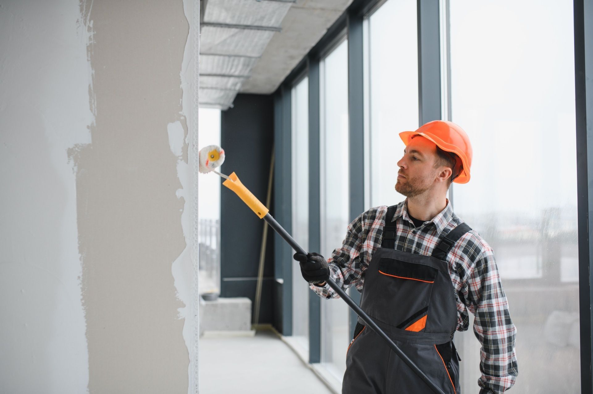Man in orange hard hat paints a wall with a roller indoors.
