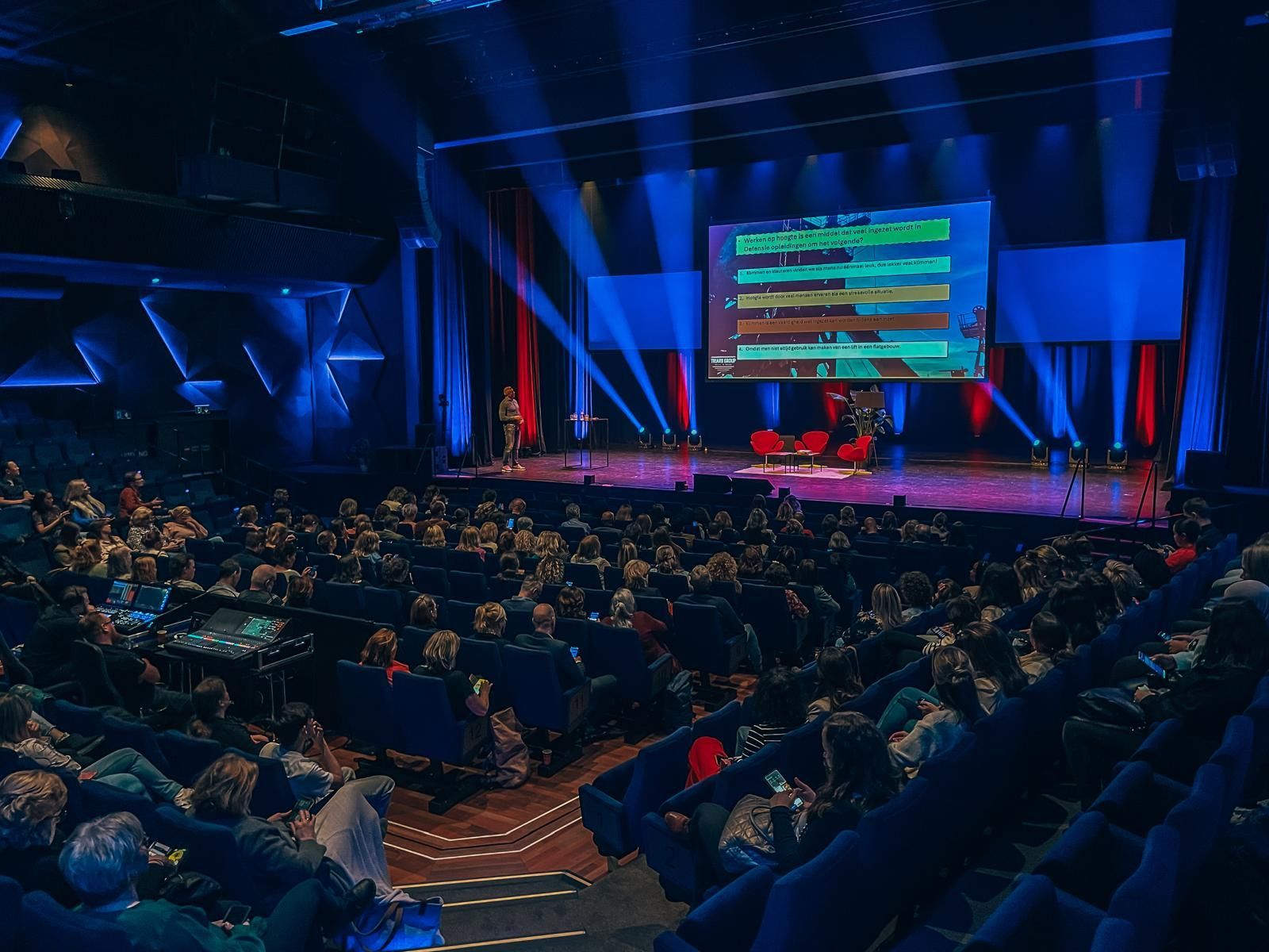 Het publiek zit in een blauw verlicht theater en kijkt naar een presentatie op een groot scherm.