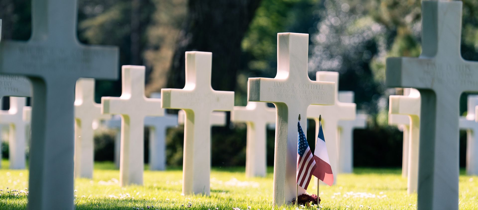 Rijen witte grafkruisen in het zonlicht, met een kleine Amerikaanse vlag op een van de graven.