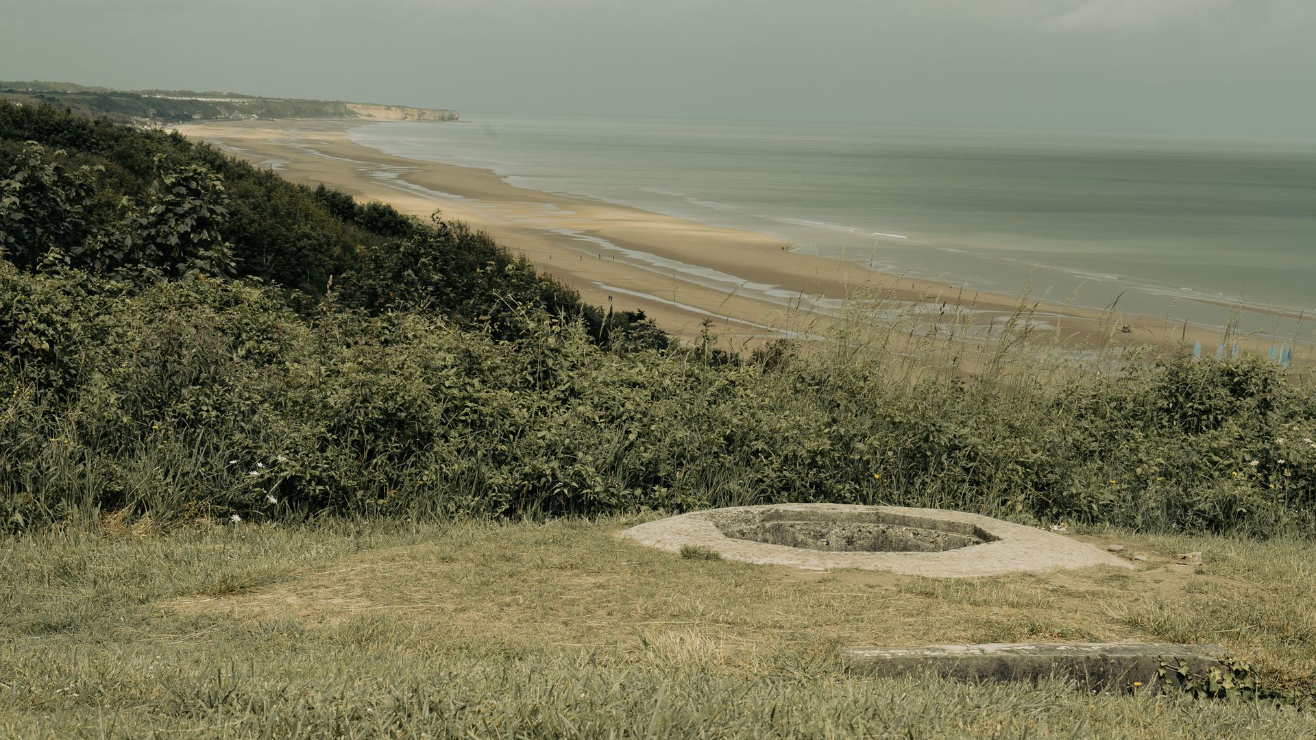 Een met gras begroeide klif aan de kust kijkt uit over een zandstrand en een kalme zee onder een nevelige hemel.