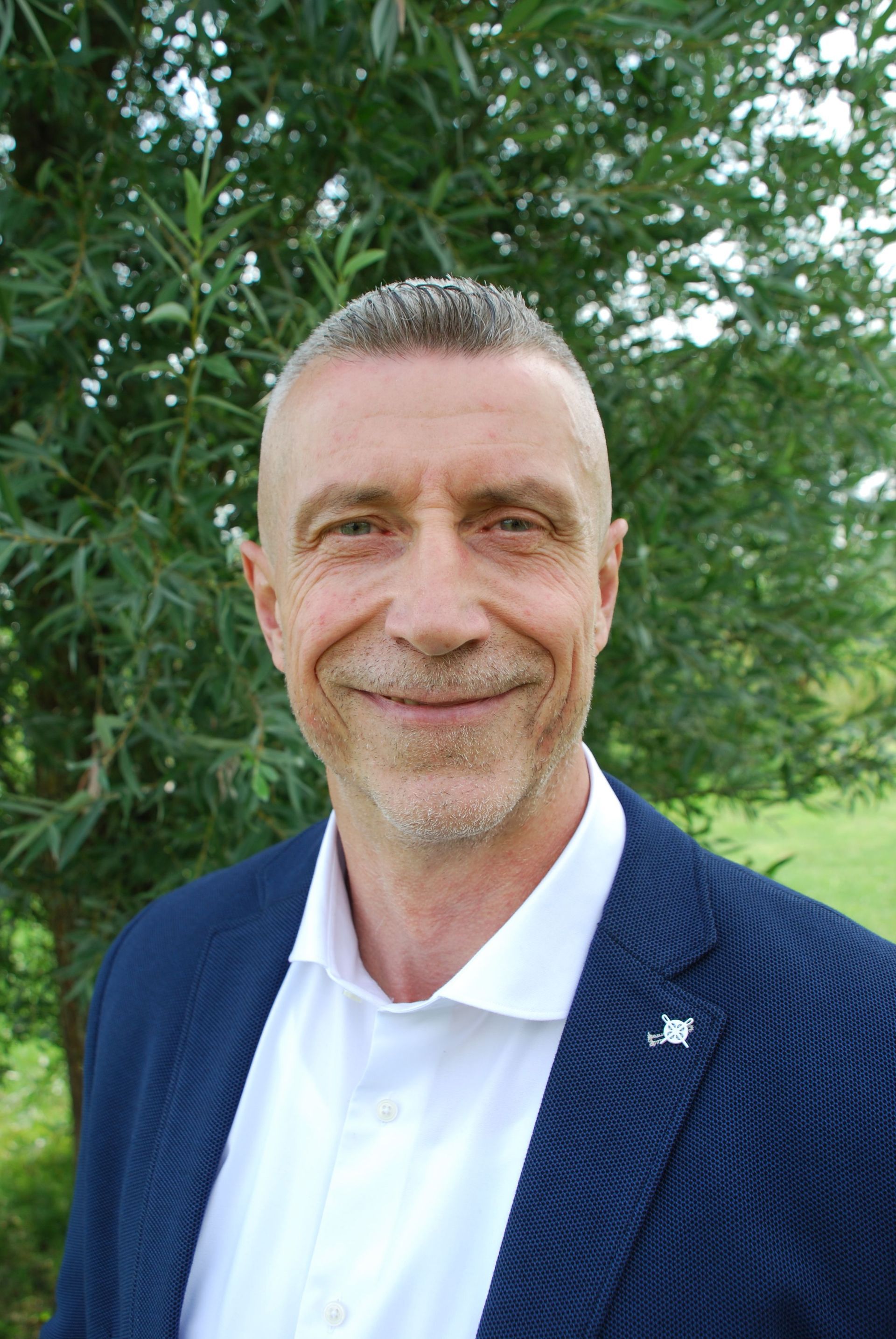 Een lachende man in een witte polo en een donkerblauwe blazer staat buiten voor groene bomen.
