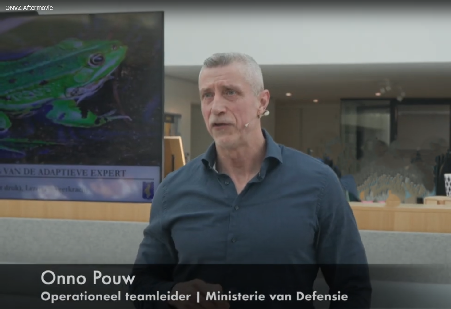 Een man staat in een moderne kantoorlobby met een kikkerposter achter hem en een Nederlandse naam op het scherm.