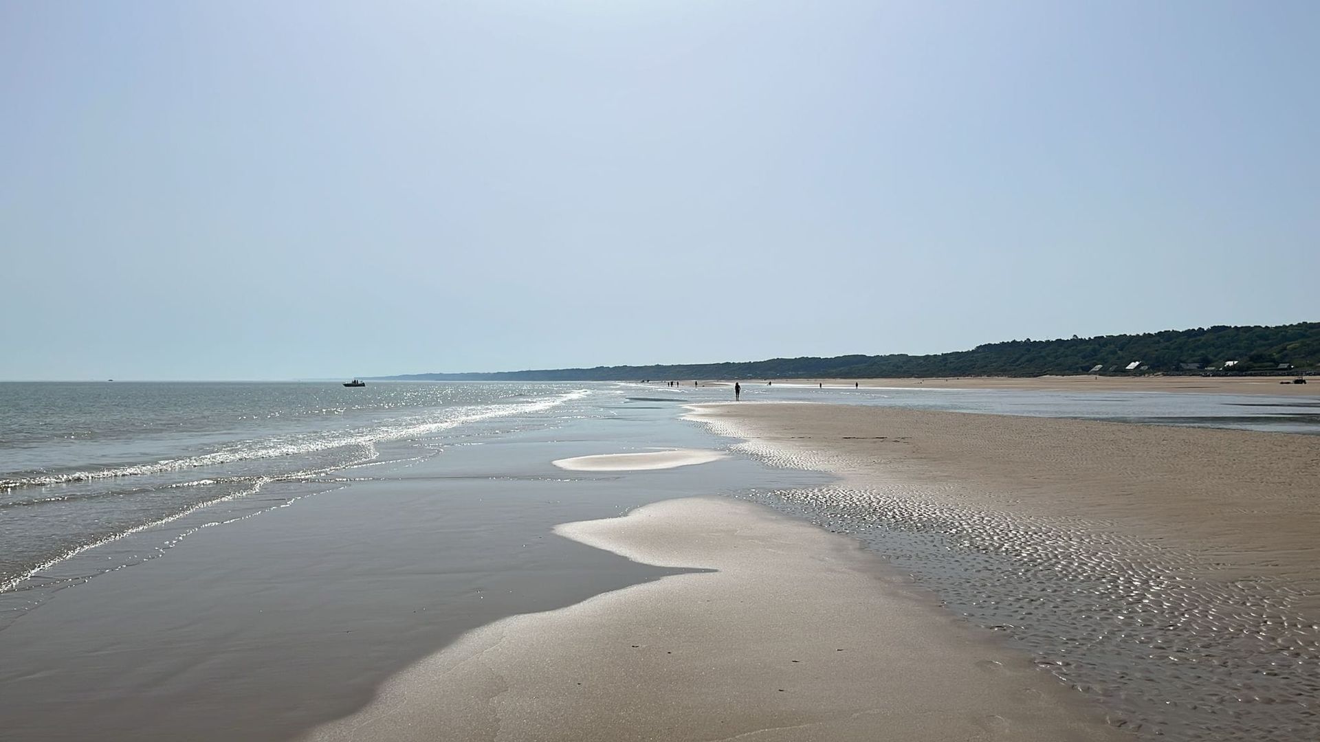 Rustig zandstrand met ondiepe golven en een verre kustlijn onder een heldere hemel.