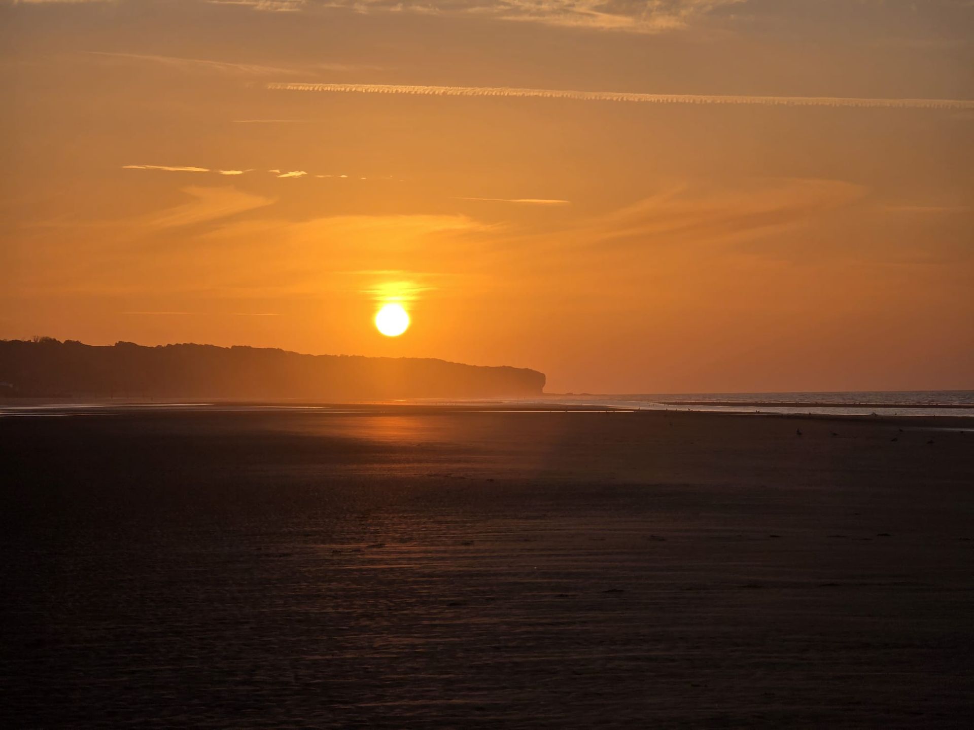 Zonsondergang boven een donker strand met een oranje lucht en kliffen in de verte.