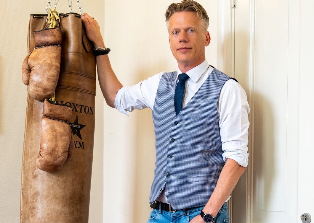 Een man staat naast een hangende bokszak in een witte kamer, gekleed in een blauw gestreept vest en stropdas.
