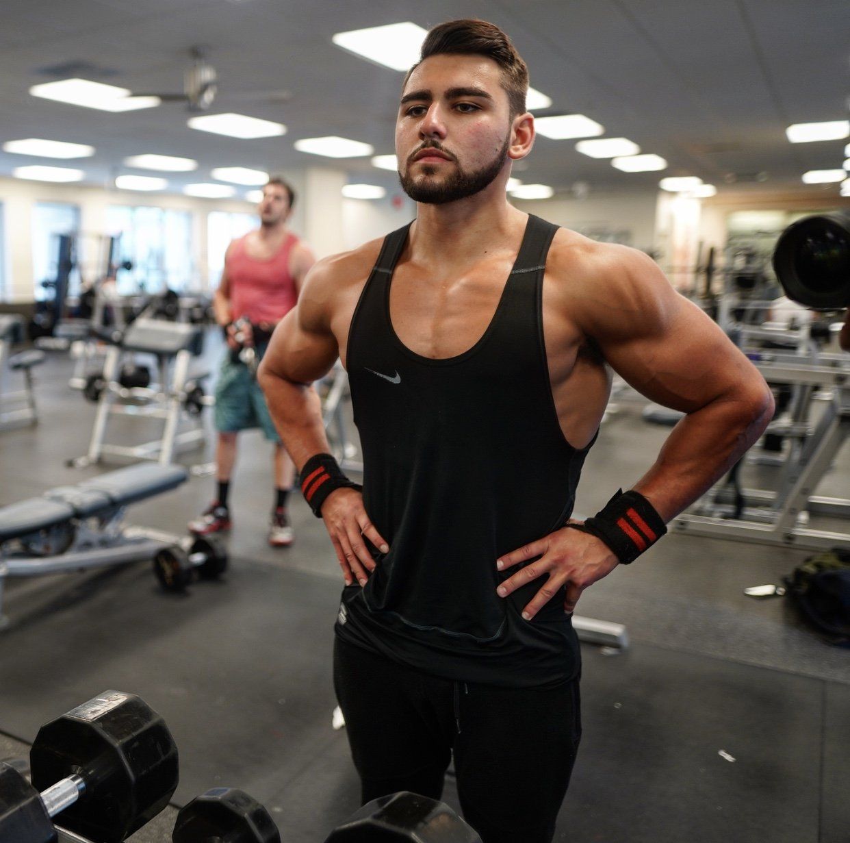 A man in a black tank top is standing in a gym with his hands on his hips