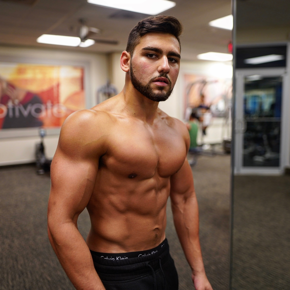 A shirtless man with a beard is standing in front of a mirror in a gym.