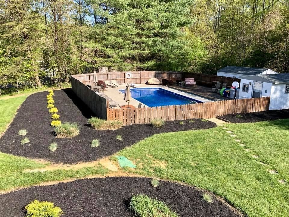 Backyard pool deck with blue pool, wooden fence, lawn, and landscaped mulch beds surrounded by trees